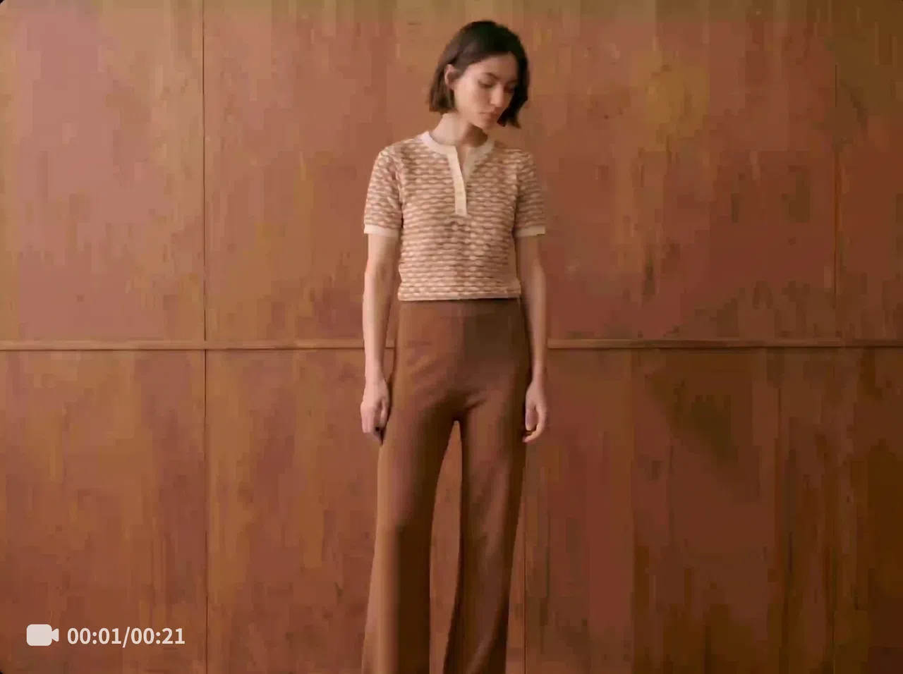 A woman in a brown-and-white top and brown trousers stands in front of a wooden wall