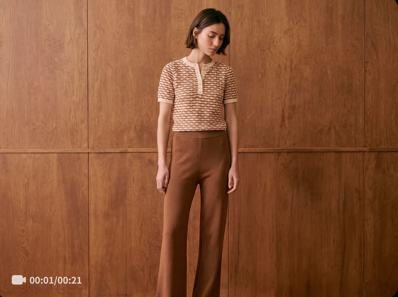 A woman in a brown-and-white top and brown trousers stands in front of a wooden wall