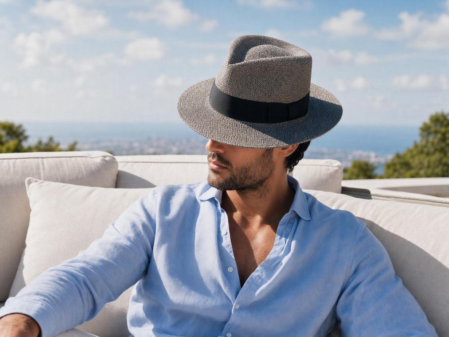 Modelo masculino con sombrero fedora de paja relajándose en un patio al aire libre con vista al mar