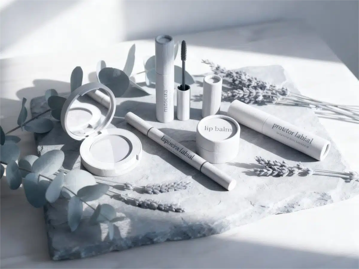 Uma coleção de produtos de maquiagem sustentáveis em tons frios e desaturados, dispostos sobre uma pedra cinza com ramos de lavanda e eucalipto sob uma luz suave e clara.