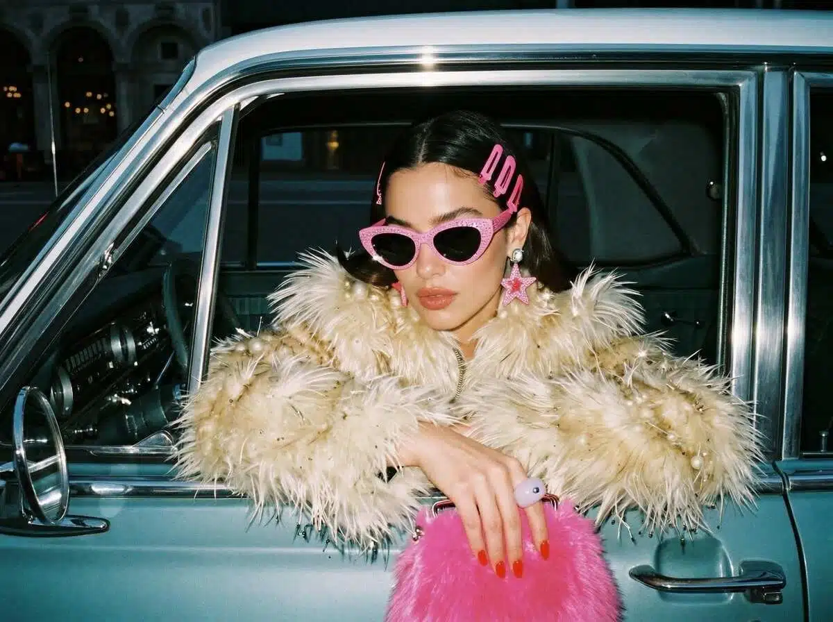 Uma mulher estilosa inclinada para fora da janela de um carro vintage azul-petróleo. Ela usa óculos escuros com strass rosa, brincos em forma de estrela e um volumoso casaco de pele sintética creme, segurando uma bolsa de pelúcia rosa vibrante.